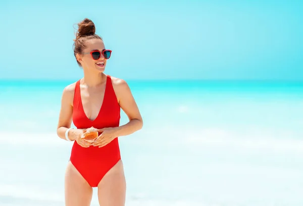 Glückliche moderne Frau im roten Badeanzug mit Muschel am Strand — Stockfoto