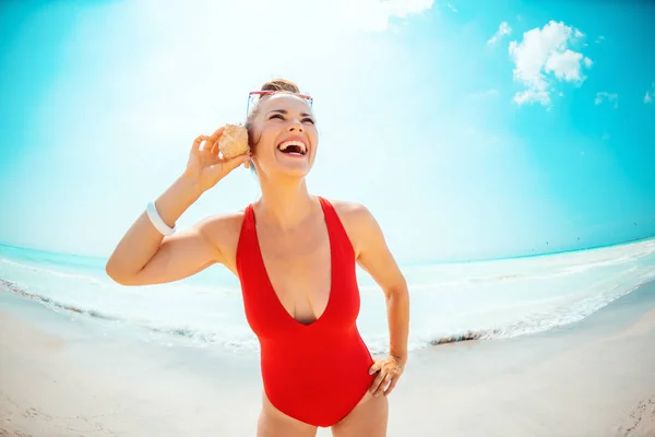 Femme souriante sur la plage à l'écoute du son de l'océan en coquille — Photo