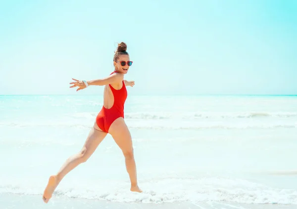 Gelukkige moderne vrouw in rode badmode op strand rennen — Stockfoto