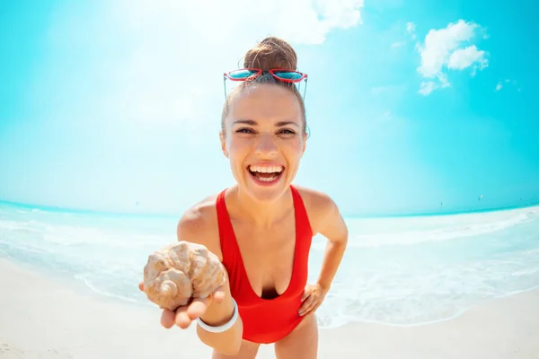 Mulher moderna feliz em roupa de banho vermelha na praia mostrando concha do mar — Fotografia de Stock