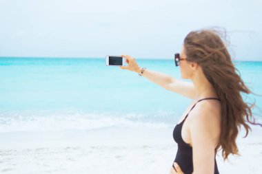 Beyaz bir plajda akıllı telefon ile fotoğraf çekmek güneş gözlüğü şık orta yaş kadın arkasından görüldü.