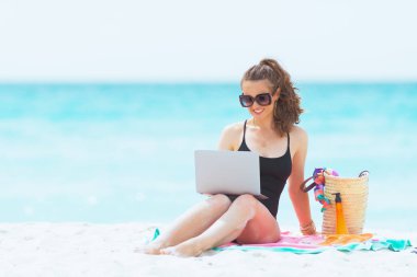 beyaz bir plajda dizüstü bilgisayar ile zarif siyah mayo uzun kıvırcık saçlı mutlu trendy orta yaş kadın.
