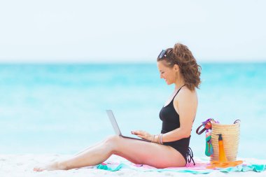 beyaz bir plajda dizüstü bilgisayar ile zarif siyah mayo uzun kıvırcık saçlı mutlu genç 40 yaşındaki kadın.