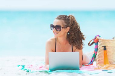 beyaz bir plajda dizüstü bilgisayar ile zarif siyah mayo uzun kıvırcık saçlı mutlu trendy orta yaş kadın.