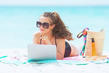 beyaz bir plajda zarif siyah mayo uzun kıvırcık saçlı pensive zarif orta yaş kadın bir dizüstü bilgisayar üzerinde araştırma yapıyor.