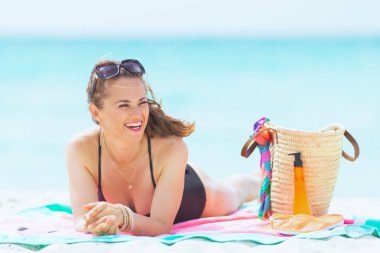 yuvarlak karpuz havlu üzerinde yatarken mesafe içine bakarak beyaz bir plajda zarif siyah mayo uzun kıvırcık saçlı modern orta yaş kadın.