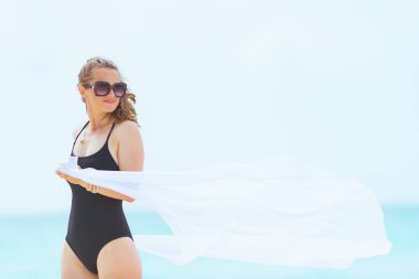 beyaz pareo tutan beyaz bir plajda zarif siyah mayo uzun kıvırcık saçlı trendy 40 yaşındaki kadın.