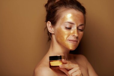 Bronz arka plan üzerinde izole yüz krem şişe tutan altın kozmetik yüz maskesi ile modern orta yaş kadın portresi.