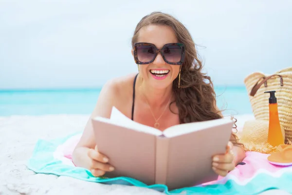 微笑优雅的中年妇女与长卷发在优雅的黑色泳衣在白色海滩阅读书的肖像 — 图库照片