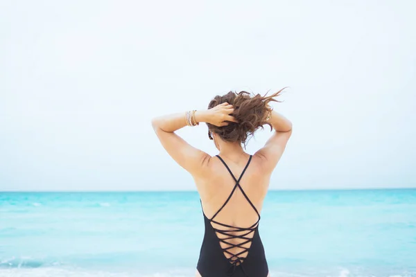 Seen from behind stylish 40 year old woman with long curly hair in elegant black swimsuit on a white beach adjusting hair.