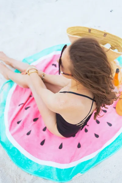 stock image Upper view of modern middle age woman with long curly hair in elegant black bathing suit on a white beach sitting on round watermelon towel.