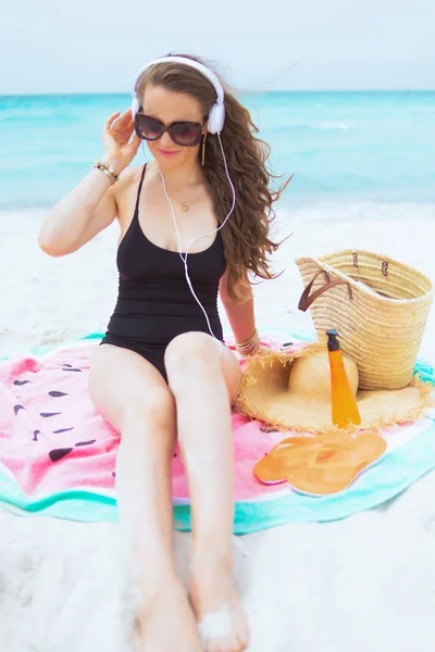 白いビーチで丸いスイカタオルに座っている間 ヘッドフォンで音楽を聴いてエレガントな黒い水着で長い巻き毛を持つ現代の40歳の女性を微笑む — ストック写真
