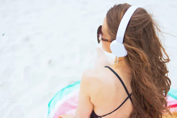 Seen from behind young middle age woman with long curly hair in elegant black swimsuit looking into the distance and listening to the music with headphones on a white beach.