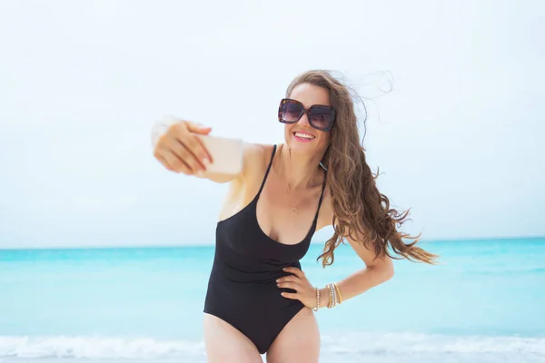 smiling young middle age woman in sunglasses taking selfie with phone on a white beach.