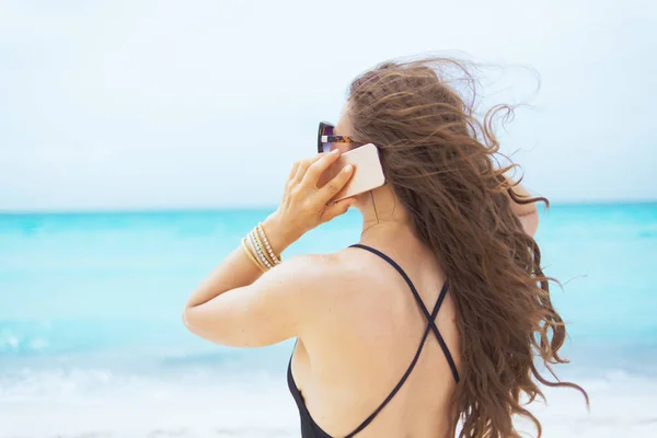 Seen from behind elegant middle age woman with long curly hair in elegant black bathing suit on a white beach speaking on a cell phone.