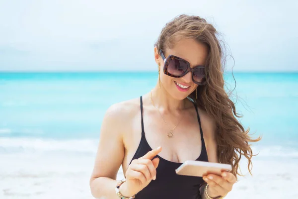 微笑的时尚中年妇女戴着太阳镜的肖像 用智能手机推动白色海滩上社交媒体的增长 — 图库照片