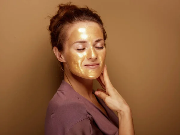 ベージュの背景に隔離された顔に触れる金色の化粧品フェイスマスクを持つ現代の40歳の女性の肖像画 — ストック写真