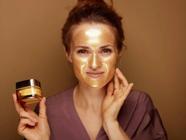 微笑优雅的40岁女子与金色化妆面罩拿着瓶脸Créme和触摸的脸在棕色背景的肖像 — 图库照片