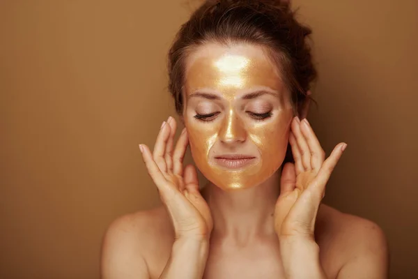 茶色の背景に顔の自己マッサージを作る黄金の化粧品のフェイスマスクを持つ現代女性の肖像画 — ストック写真