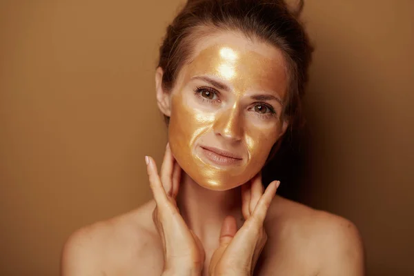茶色の背景に金色の化粧品のフェイスマスクを持つリラックスした若い女性の肖像画 — ストック写真