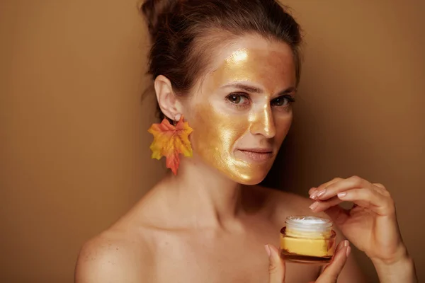 Portrait of modern 40 year old woman with golden cosmetic face mask and autumn leaf earring using cosmetic product against brown background.