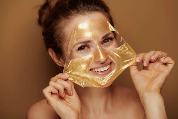 ブロンズの背景に金色の化粧品のフェイスマスクを取り除く陽気な若い女性の肖像画 — ストック写真