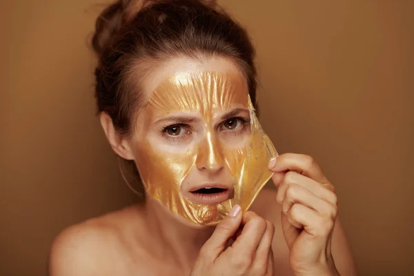 震惊优雅的40岁妇女删除金色化妆面膜隔离在米色背景的肖像 — 图库照片