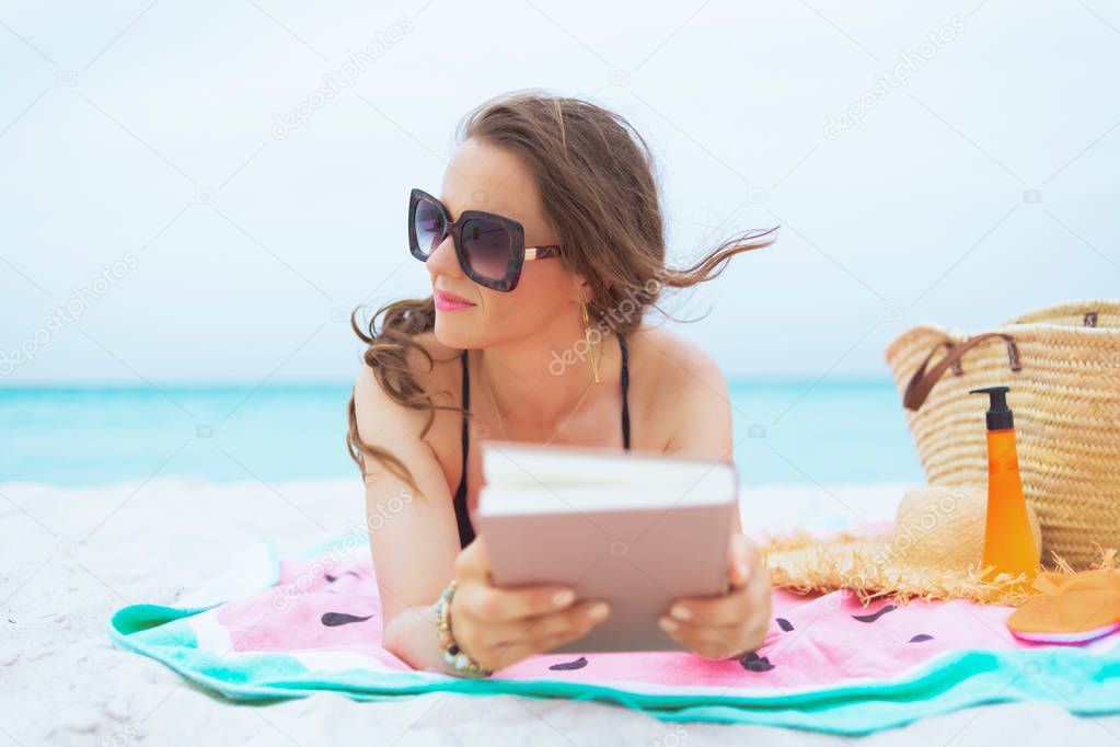 stylish 40 year old woman with long curly hair in elegant black swimsuit with book looking into the distance on a white beach.