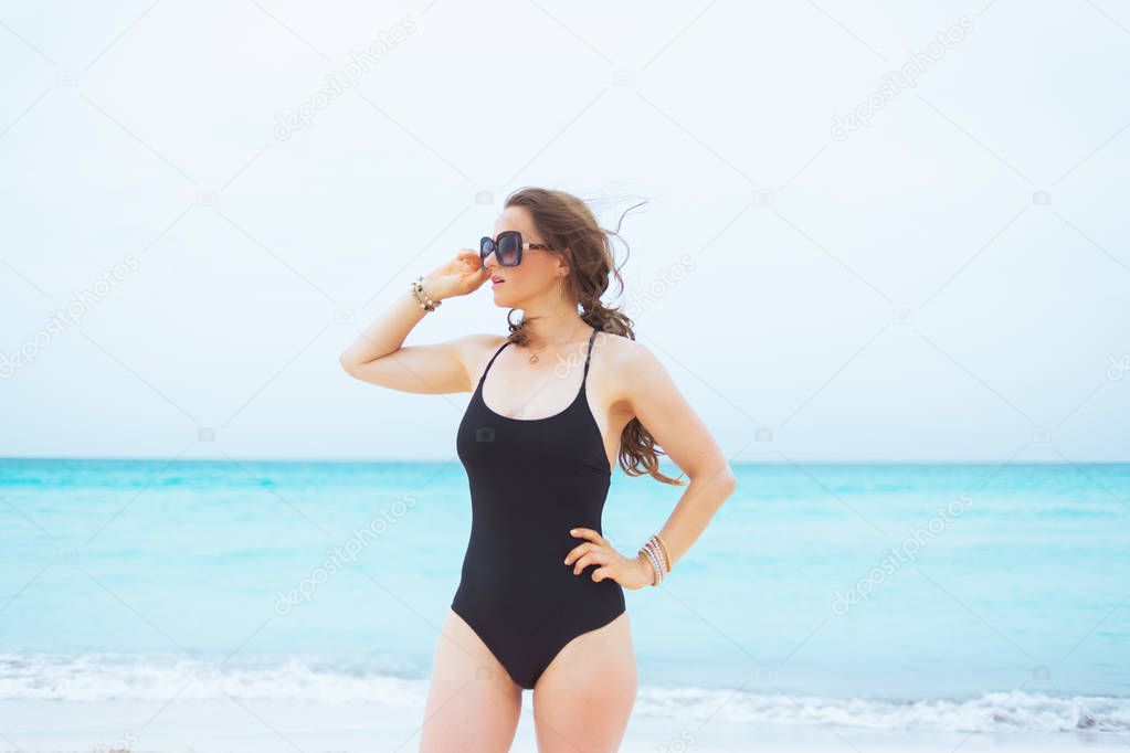 stylish middle age woman in sunglasses and with long curly hair in elegant black bathing suit on a white beach looking into the distance.