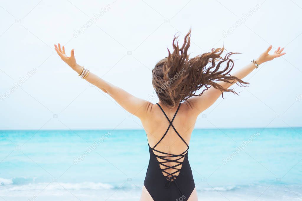 Seen from behind stylish middle age woman with long curly hair in elegant black bathing suit with raised arms rejoicing on a white beach.