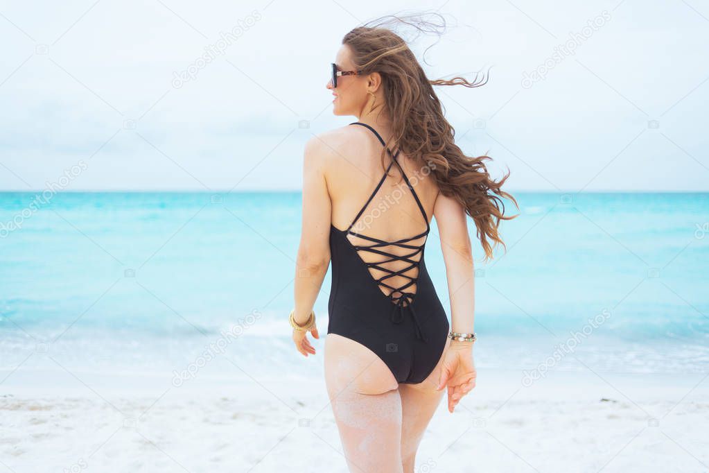 Seen from behind modern 40 year old woman with long curly hair in elegant black bathing suit on a white beach walking towards sea.