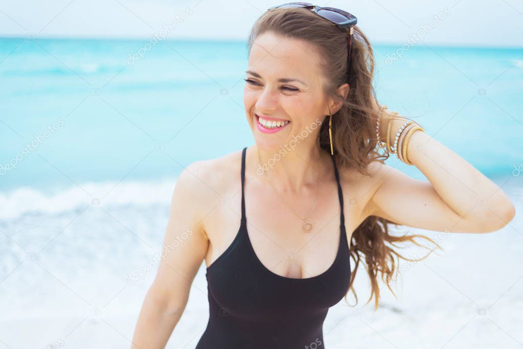 Portrait of smiling young middle age woman with long curly hair in elegant black bathing suit on a white beach looking aside.