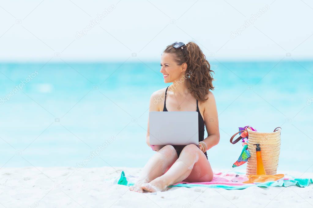 smiling trendy middle age woman with long curly hair in elegant black swimsuit on a white beach typing message on a laptop.