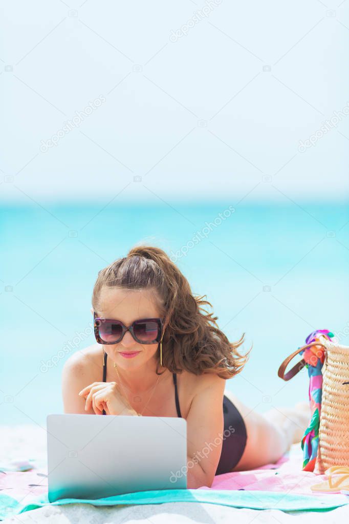 pensive elegant 40 year old woman with long curly hair in elegant black swimsuit on a white beach surfing web on a laptop.