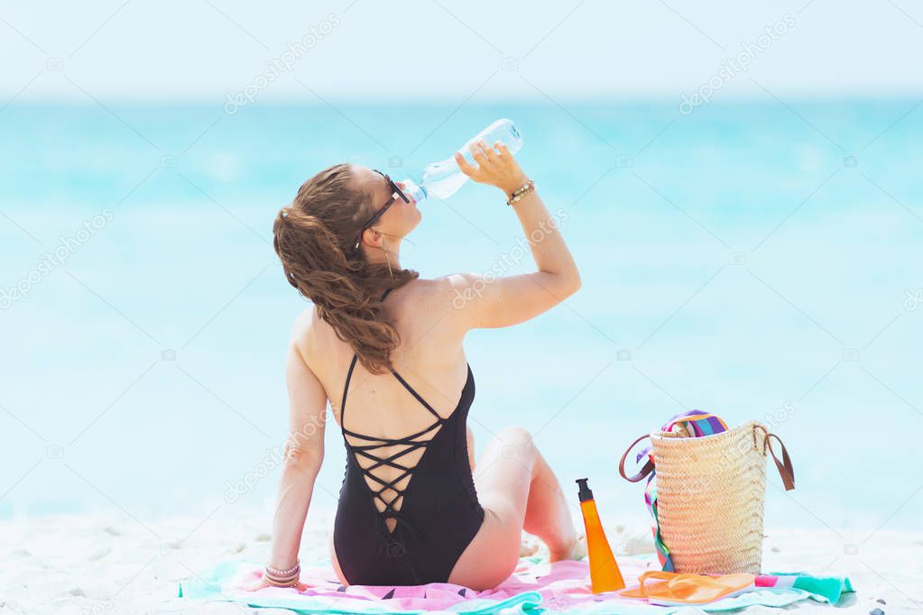 elegant 40 year old woman with long curly hair in elegant black bathing suit on a white beach drinking water from bottle while sitting on round watermelon towel.