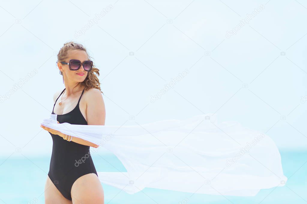 trendy 40 year old woman with long curly hair in elegant black bathing suit on a white beach holding white pareo.
