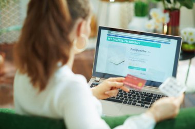 Beyaz bluzlu, kredi kartı ve boş ilaç paketi olan bir kadının dizüstü bilgisayarında online eczane kullanarak güneşli bir günde evdeki kanepede oturduğunu gördüm..
