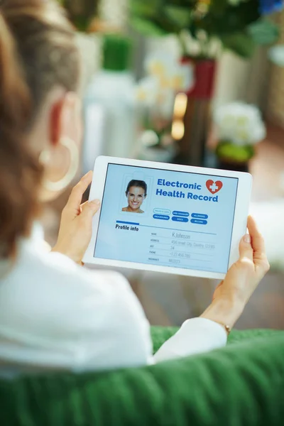 Seen from behind woman with electronic health record on a tablet PC sitting on a couch in the living room in sunny day.