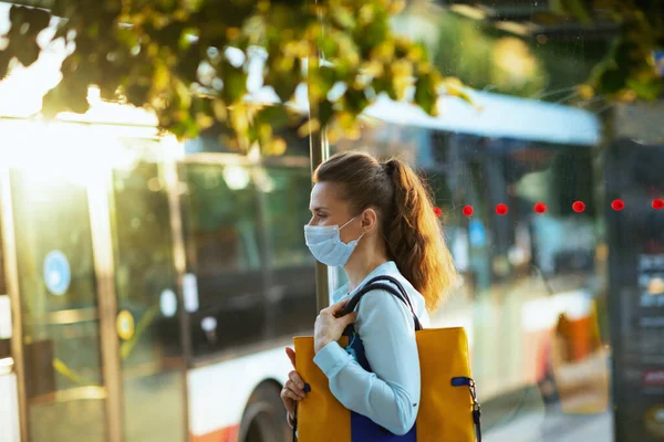 コロナウイルスのパンデミックの間の生活 優雅な女性は青いブラウスに医療用マスクとハンドバッグ街の路上でバス停での輸送を待っています — ストック写真
