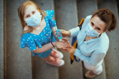 Koronavirüs salgını sırasında hayat. Tıbbi maskeli bir anne ve kızın şehir sokaklarında dezenfekte edici ile elleri dezenfekte edişinin üst görüntüsü..