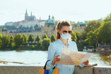 Life during coronavirus pandemic. elegant solo tourist woman in blue blouse with medical mask, sunglasses and map sightseeing outdoors on the city street. clipart