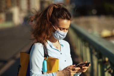 Koronavirüs salgını sırasında hayat. Medikal maskeli, çantalı ve dezenfektanlı orta yaşlı bir kadın şehirdeki akıllı telefonları kullanarak mesaj gönderiyor..