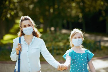 Koronavirüs salgını sırasında hayat. Sağlık maskeli mutlu anne ve çocuk şehir parkında yürüyor..