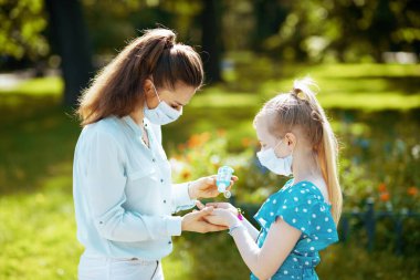 Life during coronavirus pandemic. elegant mother and daughter with medical mask disinfecting hands with sanitizer outdoors in the city park. clipart