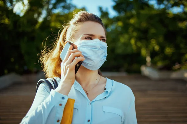 コロナウイルスのパンデミックの間の生活 青いブラウスを着た若い女性が街の屋外のスマートフォンで医療用マスクの話をしていて — ストック写真