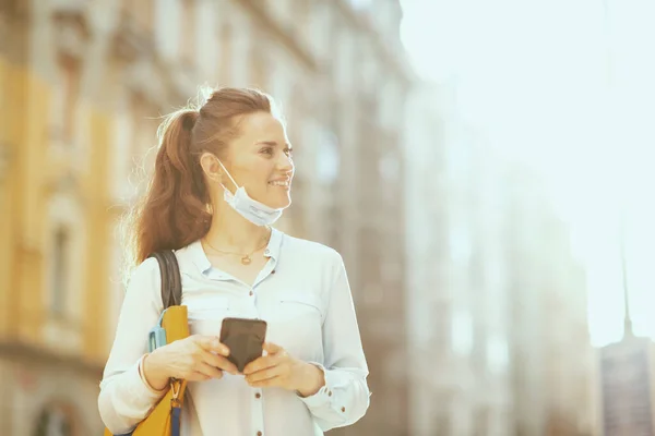 Covid 19大流行病期间的生活 在城市里 穿着蓝色衬衫 戴着医疗面具 手持手提包的快乐的年轻女性在户外使用智能手机应用程序 — 图库照片