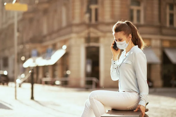 コロナウイルスのパンデミックの間の生活 都会の屋外でスマートフォンで医療用マスクの青いブラウスを着たエレガントな中年女性 — ストック写真