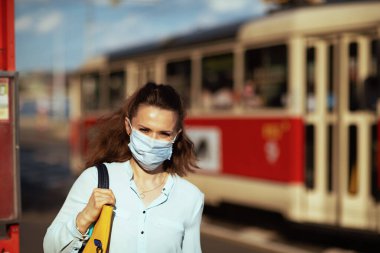 Koronavirüs salgını sırasında hayat. Şehirdeki tramvay durağında tıbbi maske ve el çantasıyla mavi bluzlu genç bir kadının portresi..
