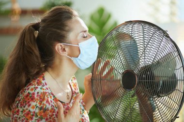 Yaz sıcağı. Çağdaş, üzgün, 40 yaşında, çiçekli bluzlu, elektrikli yelpaze ve tıbbi maskeli bir kadın güneşli bir yaz gününde modern bir evde yaz sıcağından muzdarip..