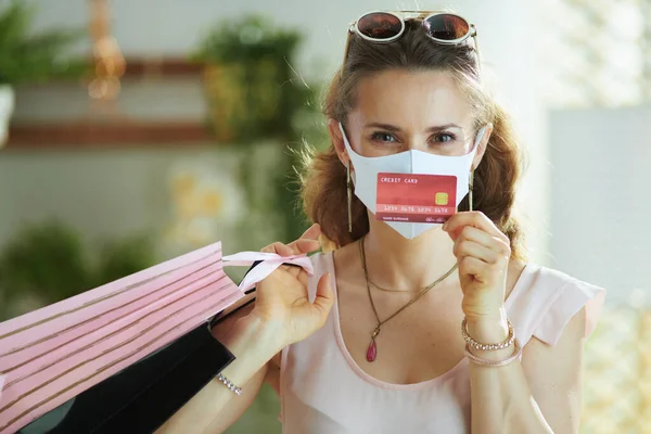 Covid 19パンデミックの間の生活 中年女性買い物客でピンクのブラウスに医療マスク クレジットカードと紙のショッピングバッグ — ストック写真